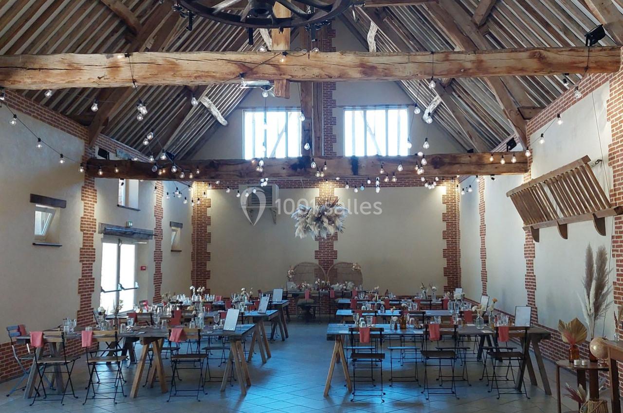 Salle de réception avec poutres apparentes, tables dressées et décorations suspendues dans un cadre rustique.