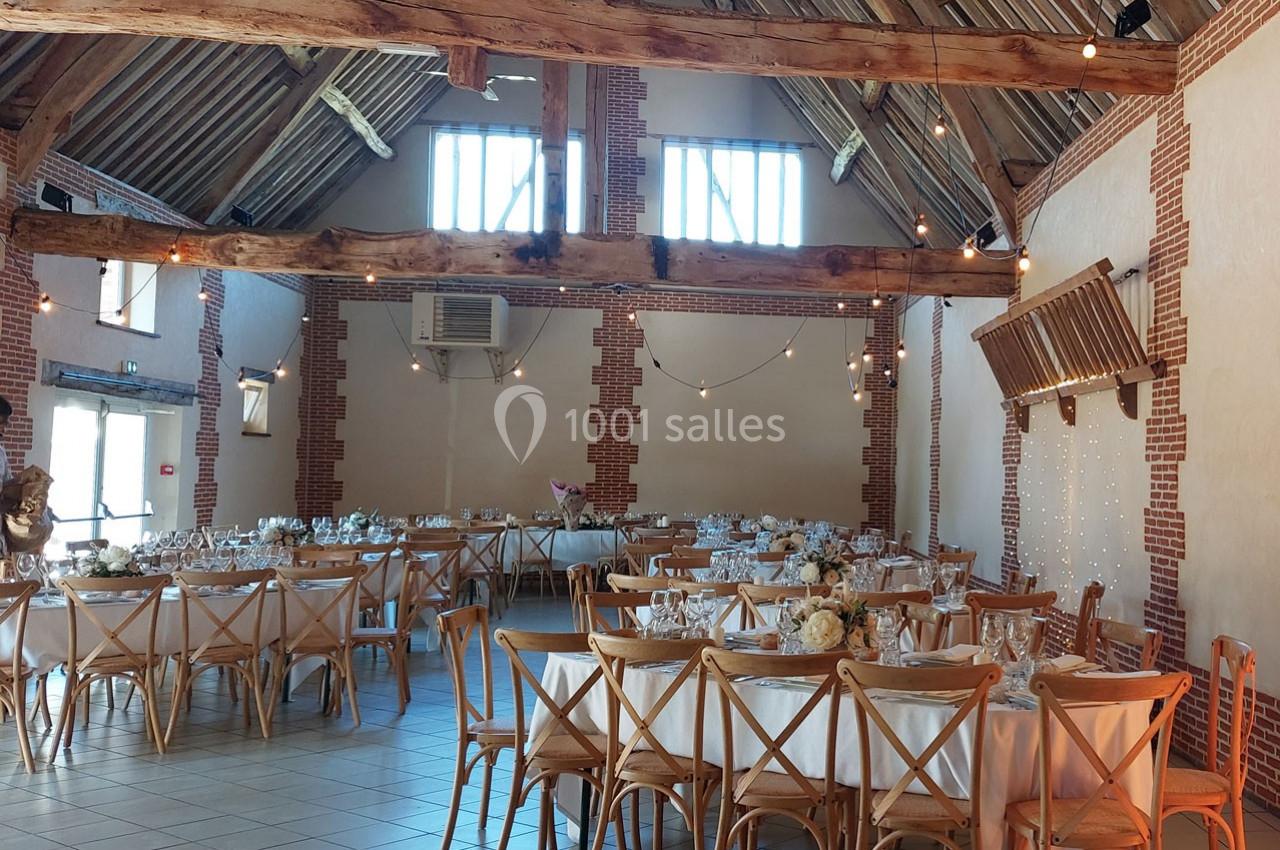 Salle de réception lumineuse avec tables dressées, chaises en bois et poutres apparentes dans un décor rustique.