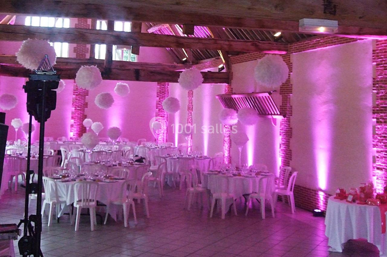 Salle de réception décorée avec des tables rondes, des nappes blanches et un éclairage rose, ornée de suspensions blanches.