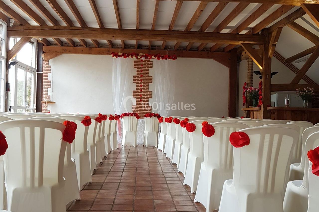 Salle décorée pour une cérémonie, avec des chaises blanches ornées de fleurs rouges alignées face à un mur en briques.
