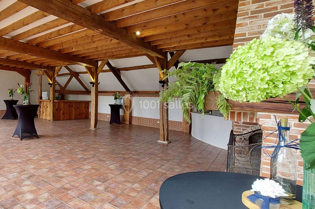 Salle avec charpente en bois apparente, sol en carrelage, tables hautes noires et décorations florales.