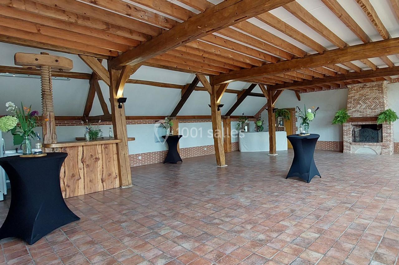 Salle avec poutres apparentes, sol en carreaux de terre cuite, tables hautes noires et cheminée en briques.