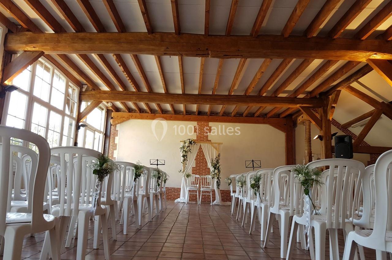 Salle décorée pour une cérémonie, avec des chaises blanches alignées et une arche ornée de fleurs au fond.