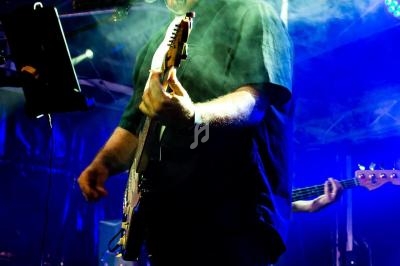 Un musicien joue de la guitare électrique et chante sur scène, éclairé par des projecteurs et entouré de fumée.