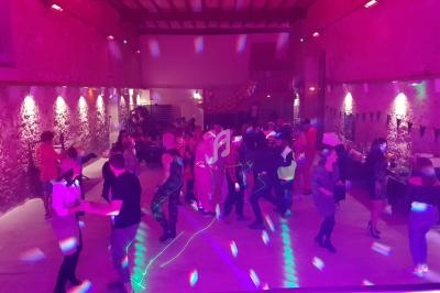 Des personnes dansent dans une salle éclairée par des lumières colorées, avec des murs en pierre et un plafond en bois.