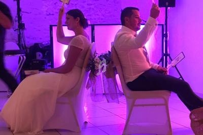 Un couple assis dos à dos lors d'un jeu de mariage, tenant des pancartes, dans une salle éclairée en violet.