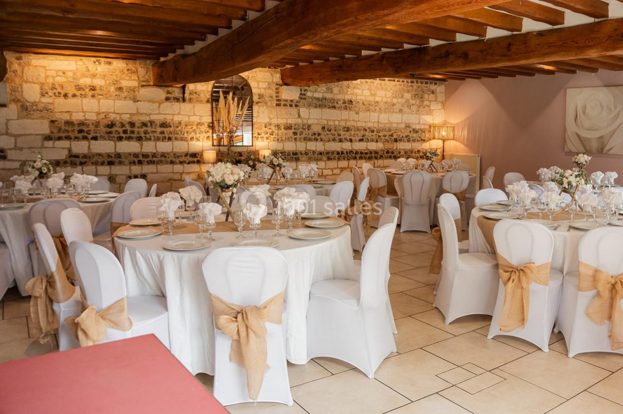 Salle de réception avec tables rondes dressées, nappes blanches, chaises décorées de nœuds beige et murs en pierre apparente.