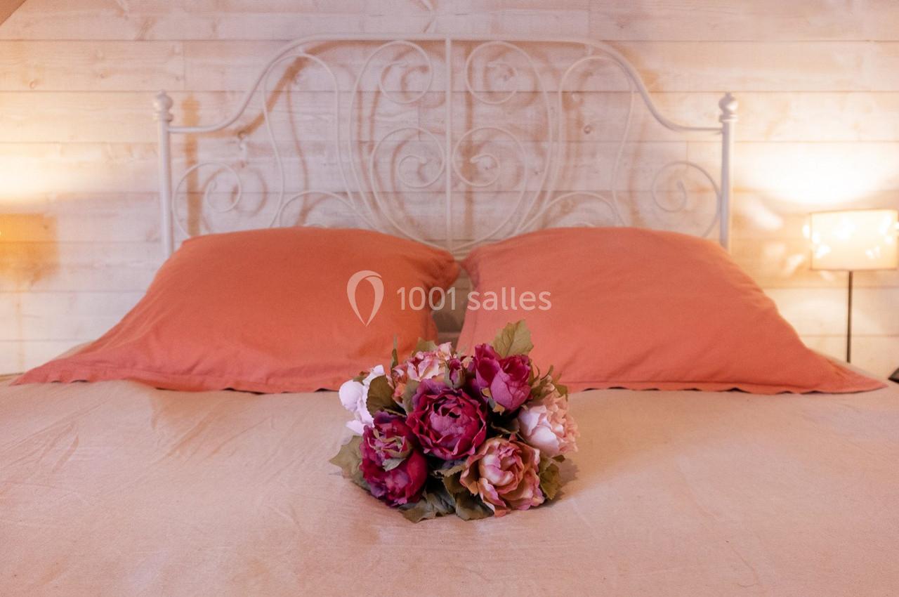 Lit avec une tête de lit en métal blanc, deux oreillers orange et un bouquet de fleurs posé sur la couverture beige.