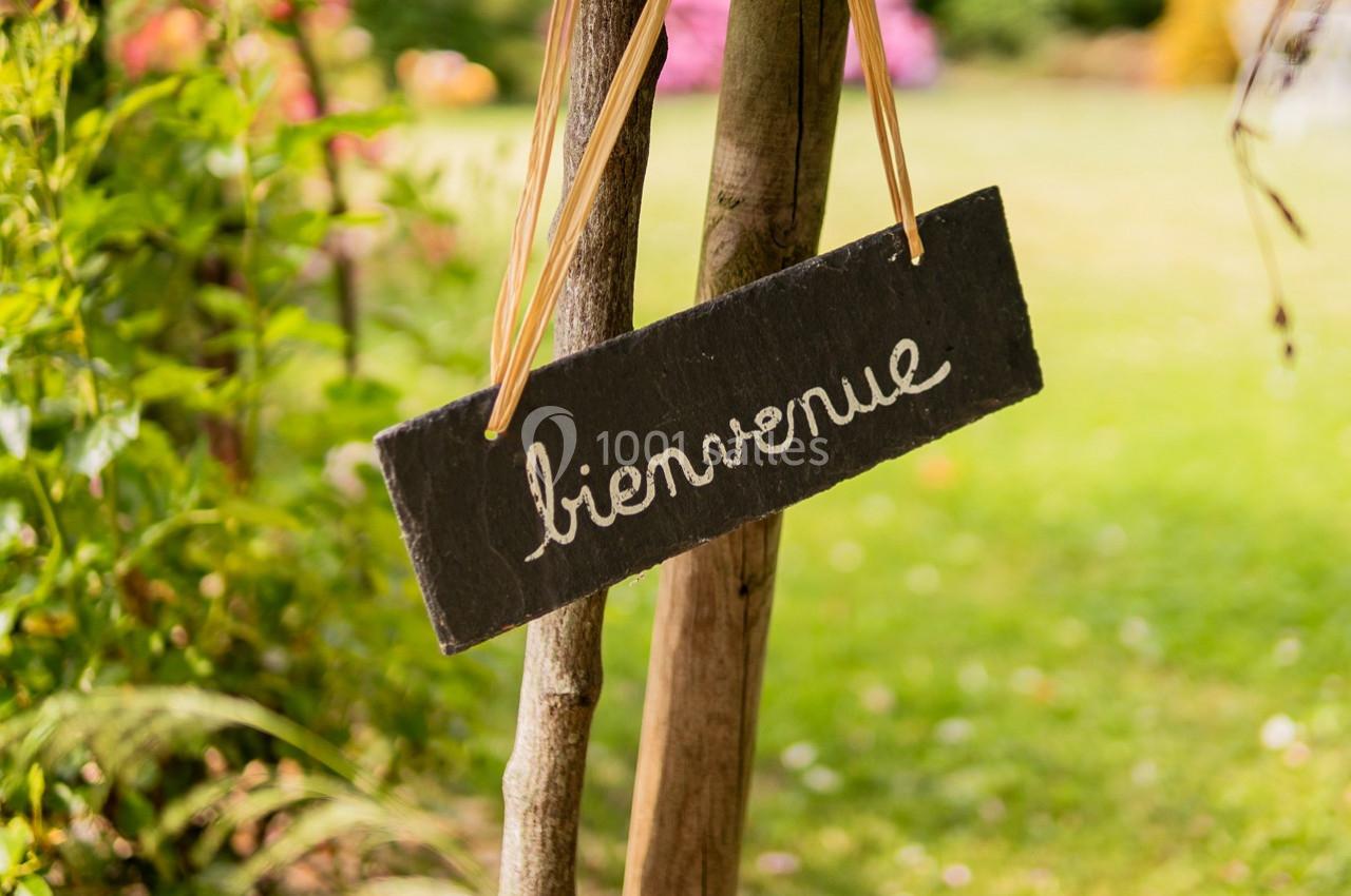Pancarte en ardoise suspendue avec l'inscription ’bienvenue’, accrochée à un poteau dans un jardin verdoyant.