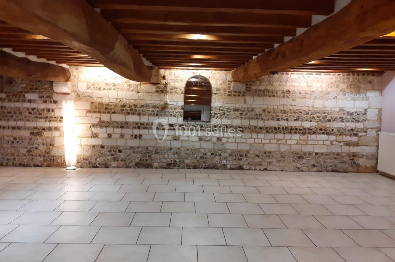 Salle vide avec murs en pierre apparente, poutres en bois au plafond et sol carrelé clair.