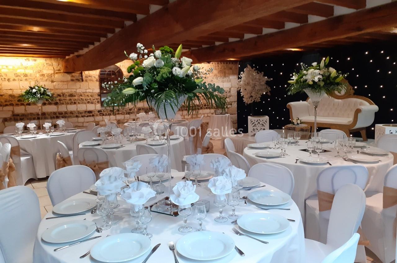 Salle de réception décorée avec des tables rondes, nappes blanches, centres de table fleuris et fauteuil en fond.