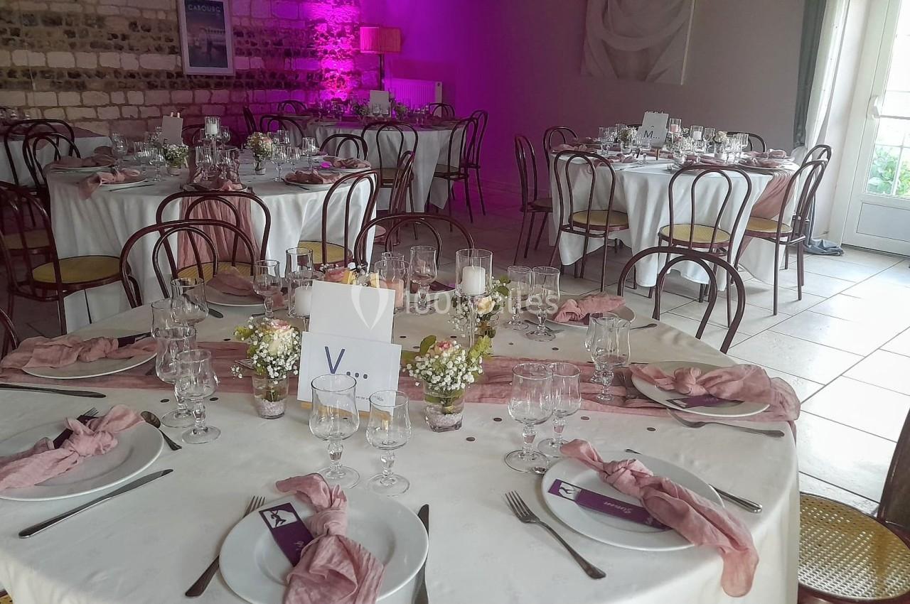 Salle de réception décorée avec des tables rondes, nappes blanches, serviettes roses et centres de table fleuris.
