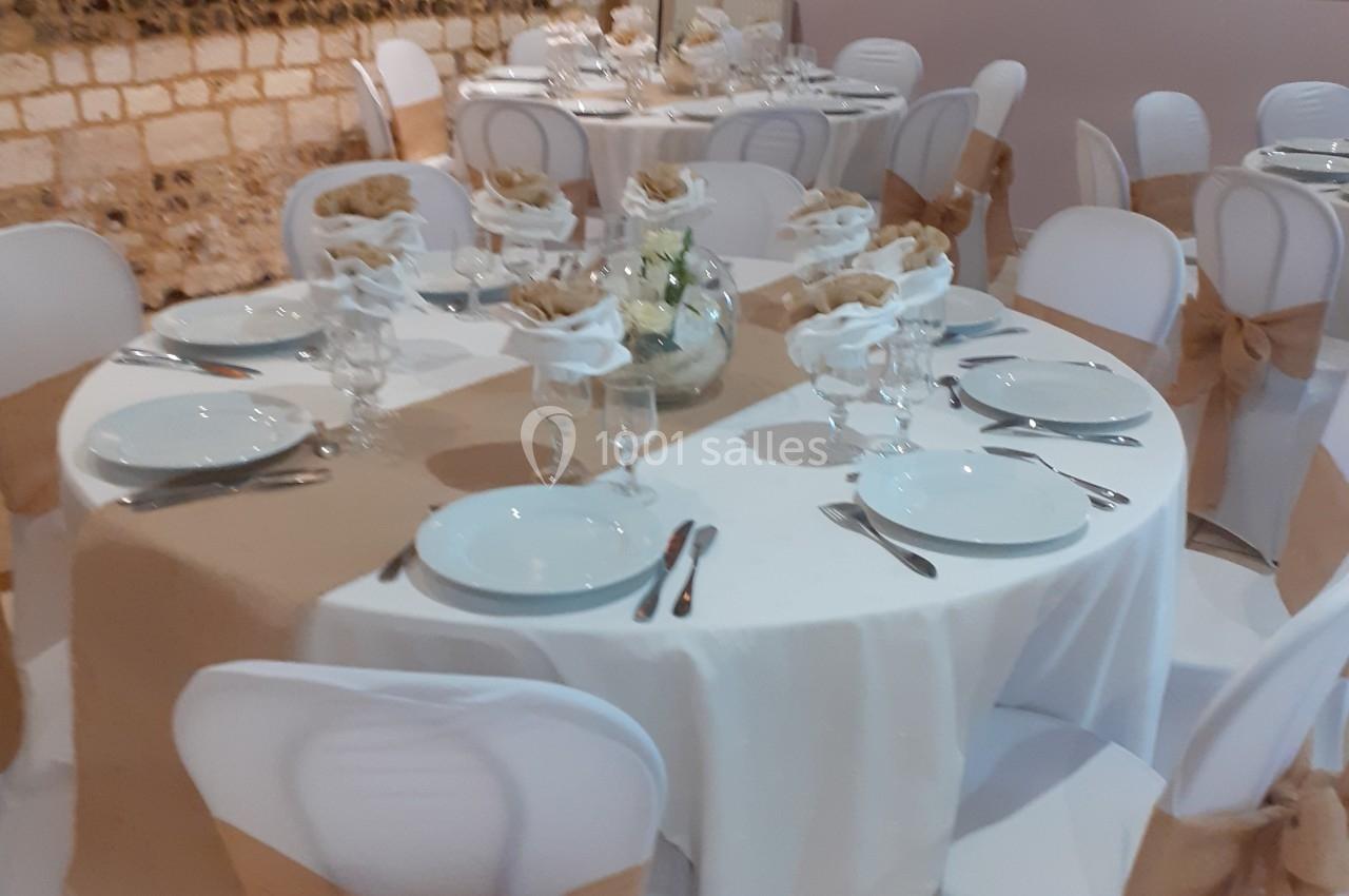 Salle décorée pour un événement avec des tables rondes dressées, nappes blanches et chaises ornées de nœuds en toile de jute.