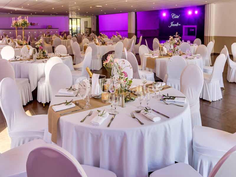 Salle de réception décorée avec des tables rondes, nappes blanches, chaises houssées et éclairage violet en arrière-plan.