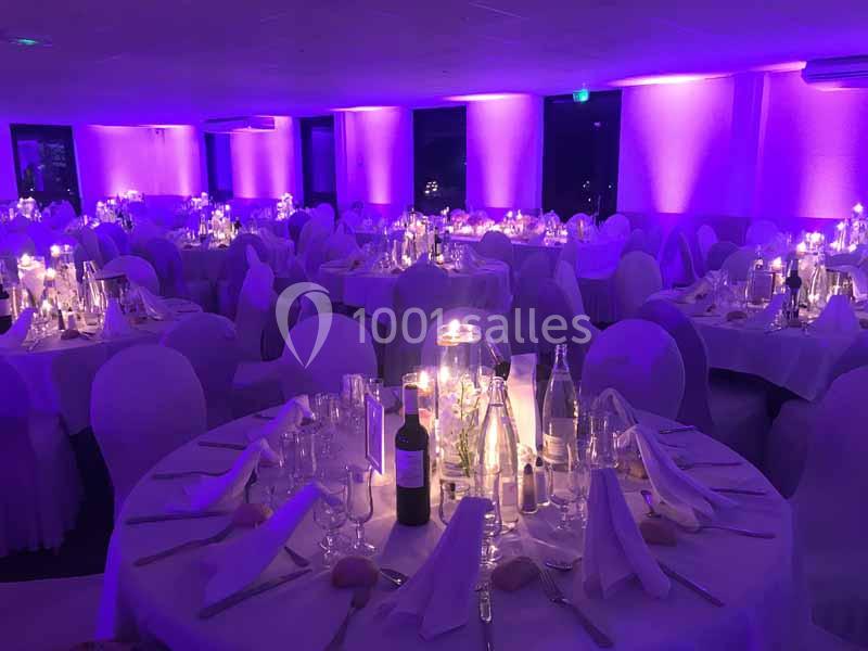 Salle de réception décorée avec des tables rondes, nappes blanches, bougies et éclairage violet tamisé.