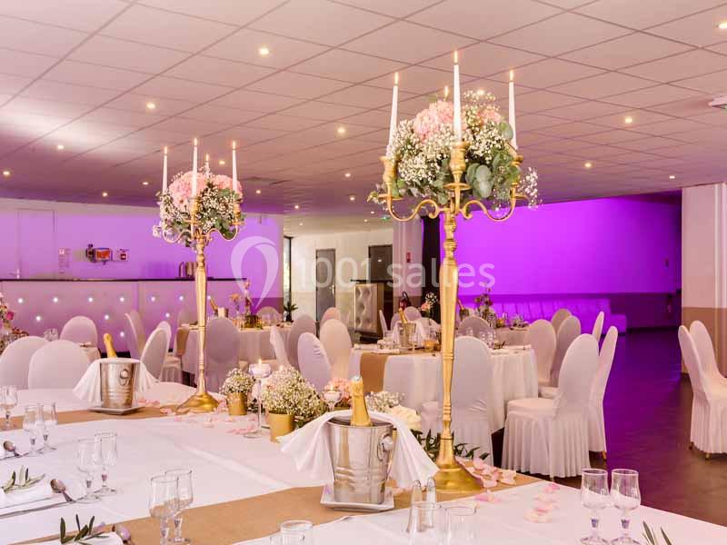 Salle de réception décorée avec des chandeliers dorés, des fleurs roses et blanches, et des tables dressées élégamment.