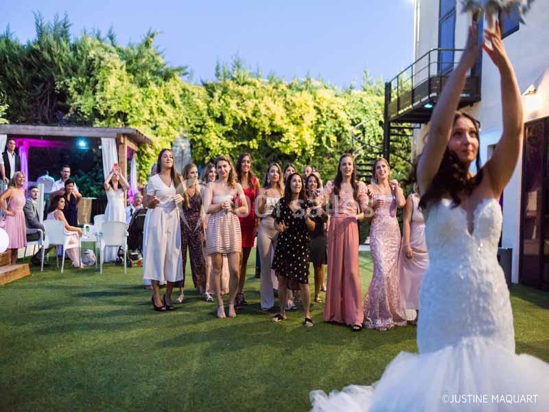Une mariée lance son bouquet à un groupe de femmes rassemblées dans un jardin lors d'une réception en soirée.
