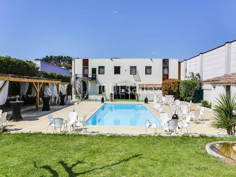 Piscine extérieure entourée de chaises blanches, pergola en bois et bâtiment moderne sous un ciel bleu.