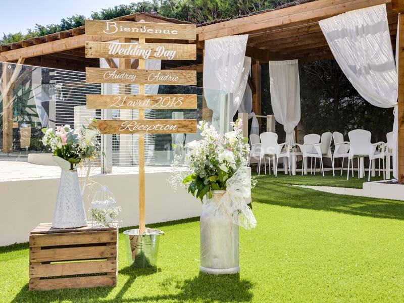 Panneau en bois indiquant un mariage, entouré de fleurs, sur une pelouse devant une terrasse décorée.