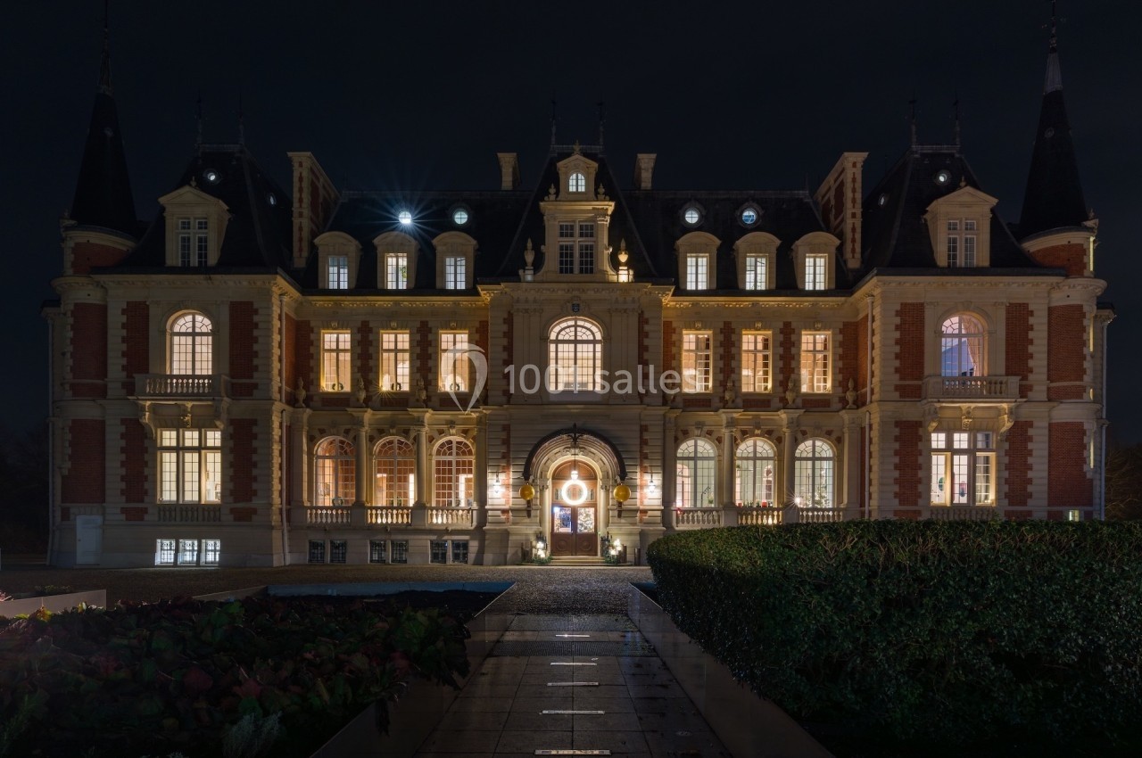 Façade d'un château illuminé de nuit, avec des fenêtres éclairées et un chemin menant à l'entrée principale.