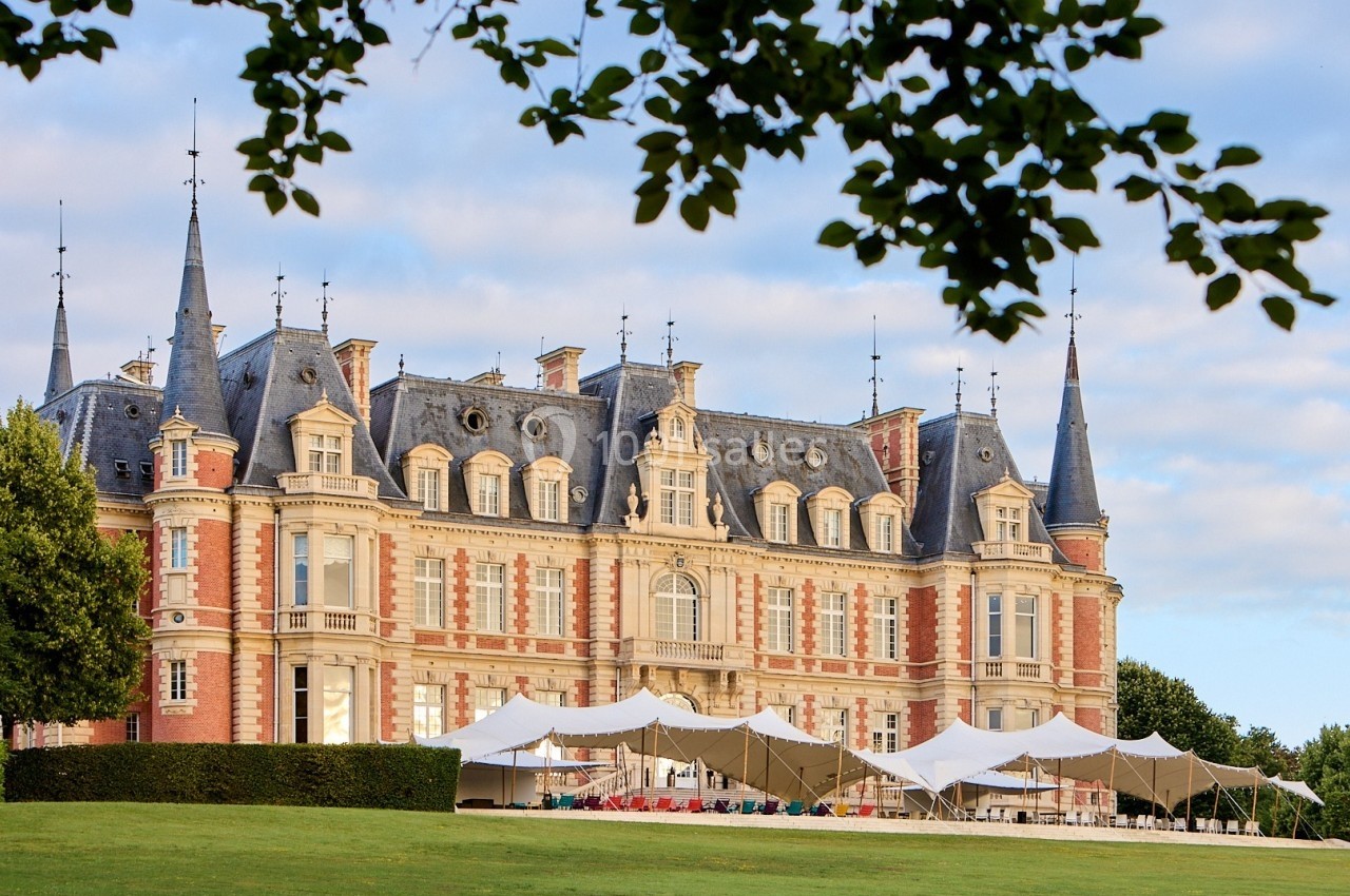 Façade d'un château en pierre et briques rouges avec des tentes blanches installées sur la pelouse devant.