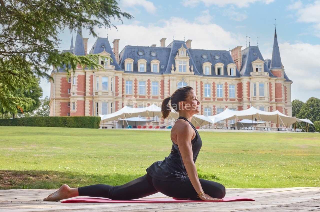 Femme pratiquant une posture de yoga sur une terrasse en bois devant un château entouré de verdure.