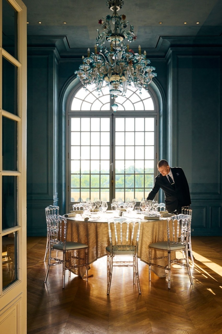 Un serveur ajuste une table élégamment dressée dans une salle lumineuse avec un grand lustre et des fenêtres cintrées.