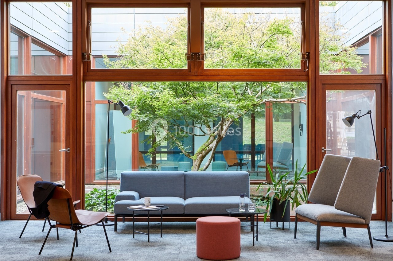 Salon moderne avec canapés, fauteuils et table basse, donnant sur un jardin arboré visible à travers de grandes baies…