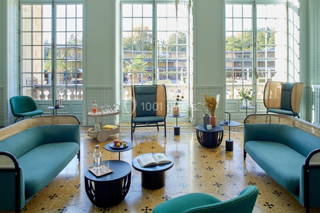 Salon lumineux avec mobilier moderne, canapés verts, tables basses et grandes fenêtres donnant sur un extérieur arboré.