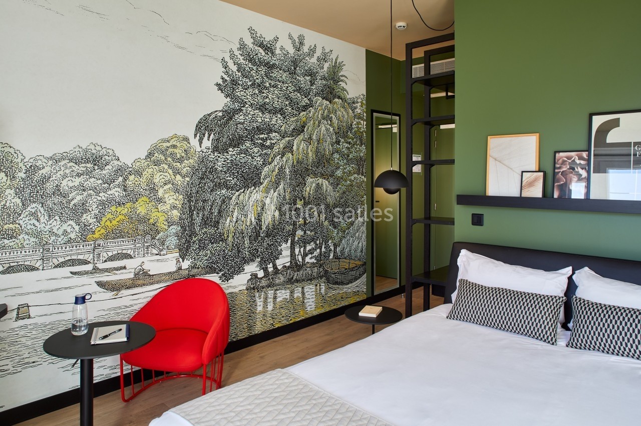 Chambre moderne avec mur vert, fresque murale de paysage, fauteuil rouge et lit avec coussins géométriques.