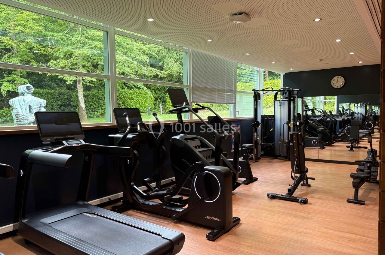 Salle de sport lumineuse avec tapis de course, vélos elliptiques, machines de musculation et miroir mural.