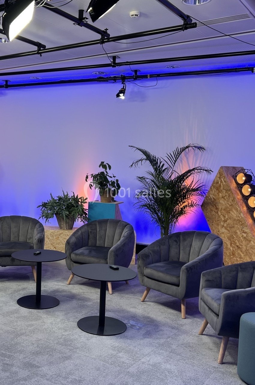 Espace de discussion moderne avec fauteuils gris, tables basses noires, plantes et éclairage coloré en arrière-plan.