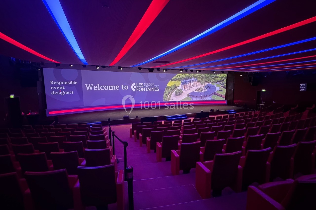 Salle de conférence vide avec sièges rouges, grand écran affichant ’Welcome to Les Fontaines’ et éclairage coloré au plafond.