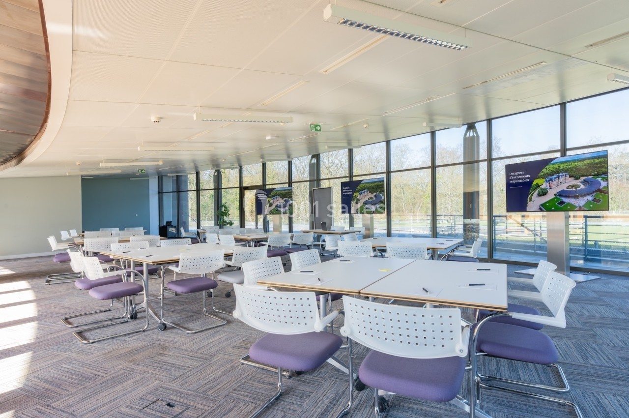 Salle de réunion lumineuse avec des tables blanches, des chaises violettes et des écrans affichant des présentations.