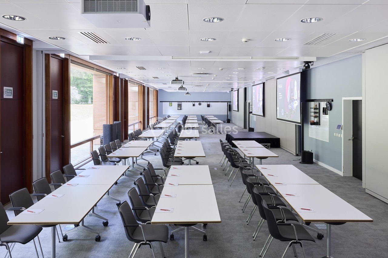 Salle de conférence lumineuse avec des tables alignées, des chaises noires et des écrans de projection au mur.