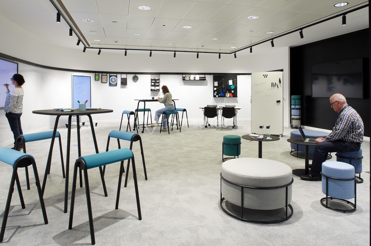 Salle moderne avec mobilier modulable, personnes travaillant sur des tables hautes et des écrans interactifs.