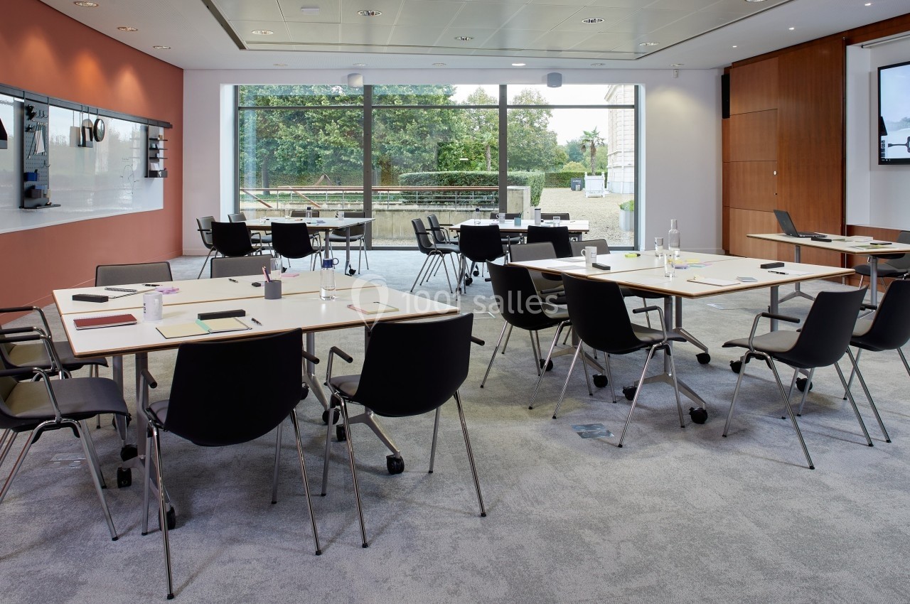 Salle de réunion moderne avec tables disposées en îlots, chaises noires et grande baie vitrée donnant sur un espace…