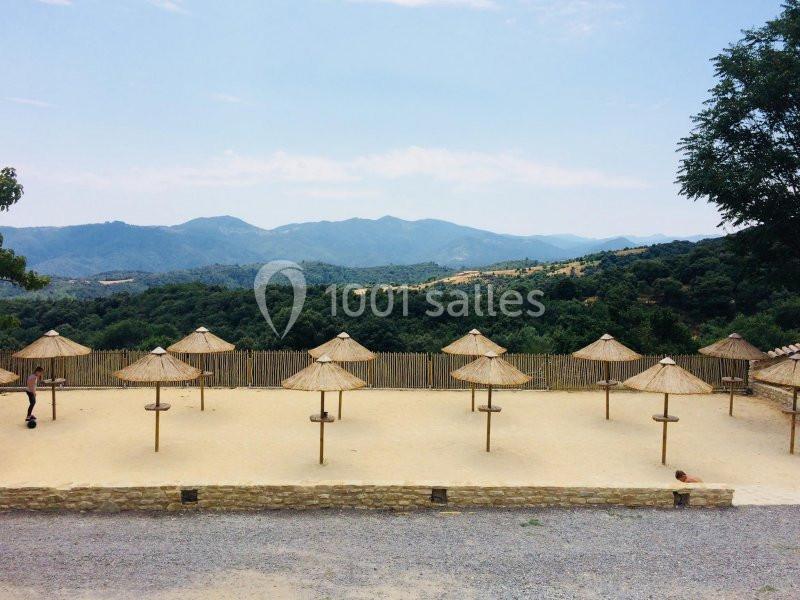 Parasols en paille alignés sur une plage de sable avec une vue sur des collines verdoyantes à l'arrière-plan.