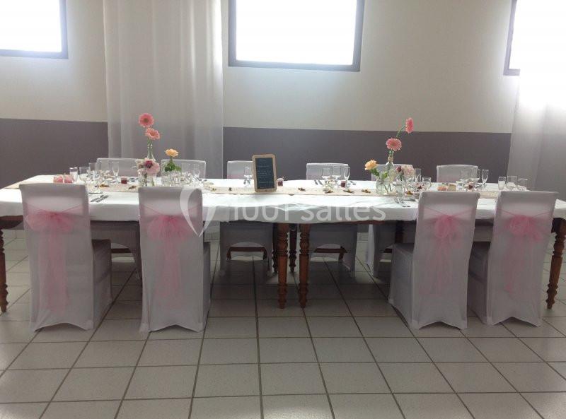 Table décorée pour un événement, avec nappes blanches, chaises ornées de nœuds roses et fleurs en centre de table.