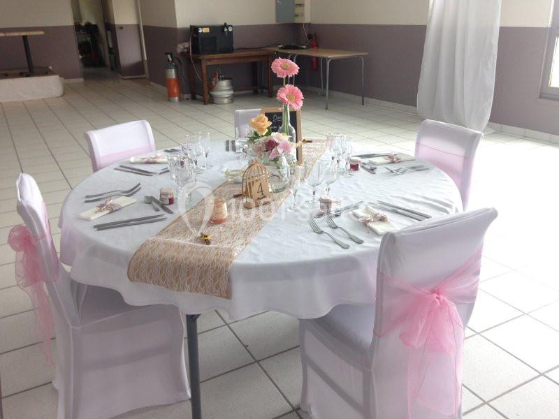 Table ronde décorée pour un événement, avec nappes blanches, chaises ornées de rubans roses et centre de table floral.