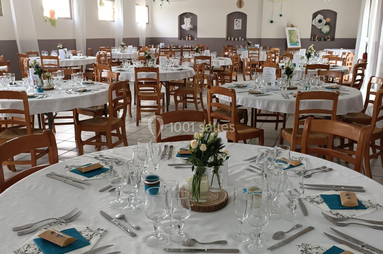 Salle de réception décorée avec des tables rondes dressées, nappes blanches, serviettes bleues et centres de table floraux.