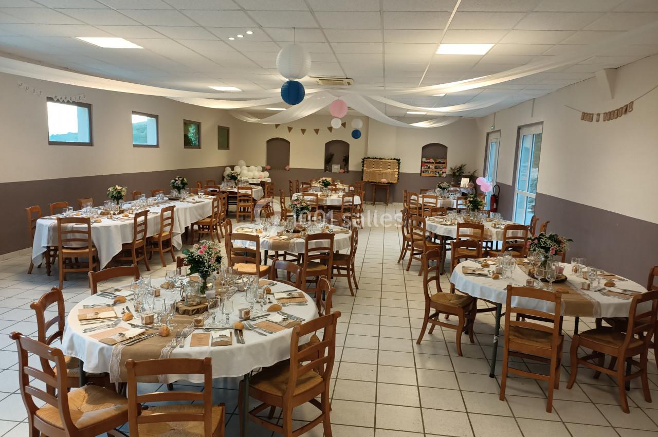 Salle décorée pour un événement avec tables rondes dressées, chaises en bois et guirlandes suspendues au plafond.
