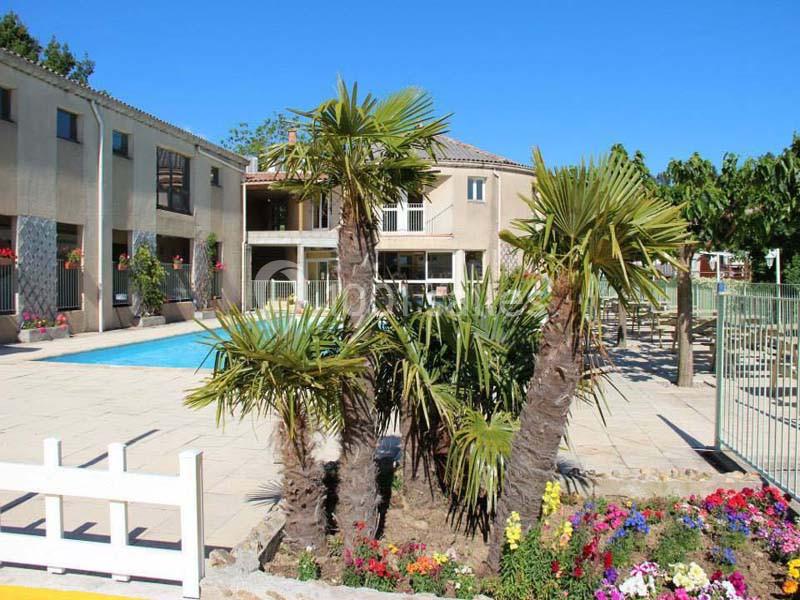 Cour intérieure avec piscine, palmiers, fleurs colorées et bâtiments en arrière-plan sous un ciel dégagé.