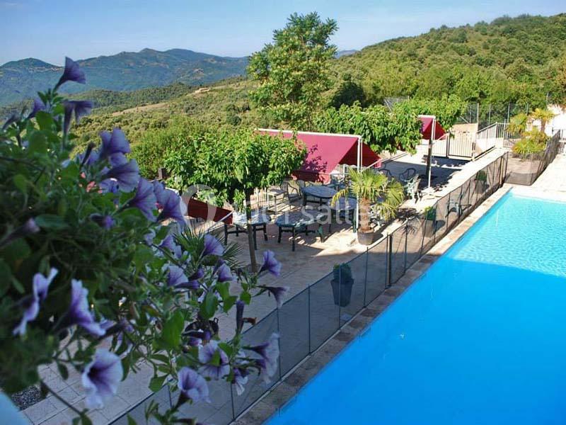 Terrasse ombragée avec tables et chaises près d'une piscine, entourée de végétation et vue sur des collines verdoyantes.