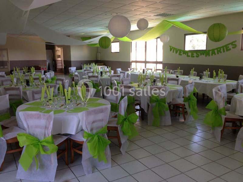 Salle décorée pour un mariage avec des tables rondes, nappes blanches, chaises ornées de nœuds verts et lanternes suspendues.