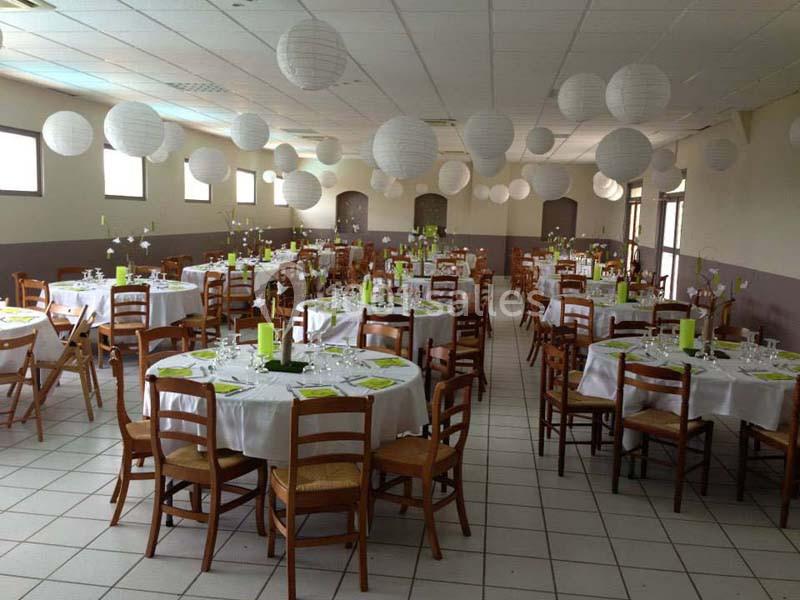 Salle de réception décorée avec des tables rondes, nappes blanches, chaises en bois et lanternes suspendues.