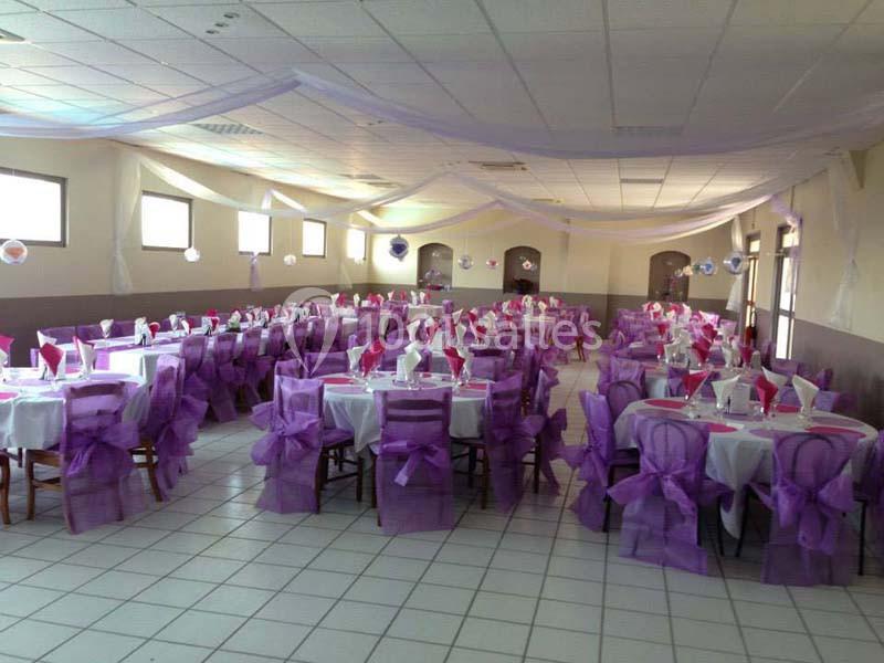 Salle de réception décorée avec des nappes blanches, des chaises ornées de nœuds violets et des guirlandes suspendues.