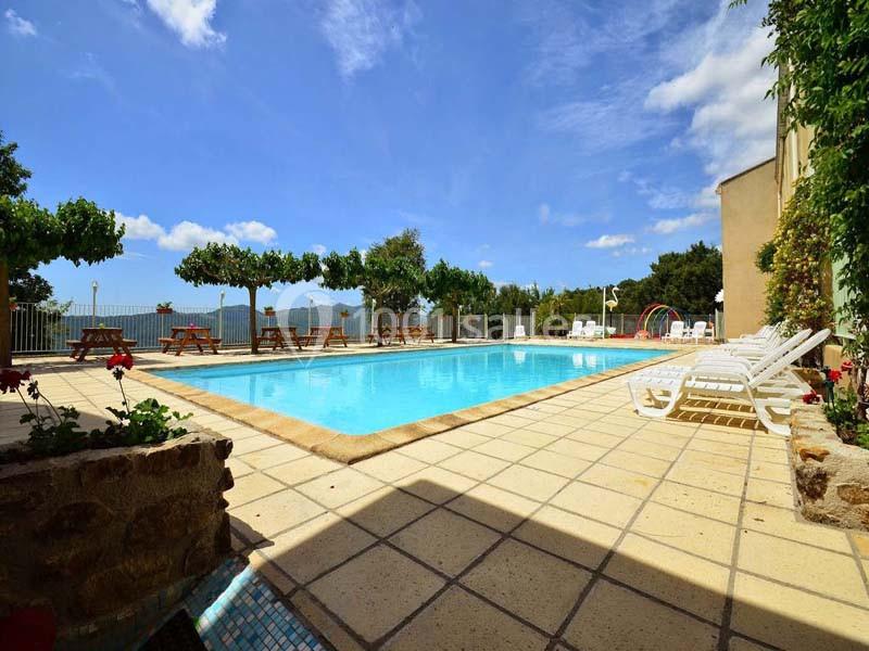 Piscine extérieure entourée de chaises longues, tables en bois et arbres, avec vue dégagée sur un paysage ensoleillé.