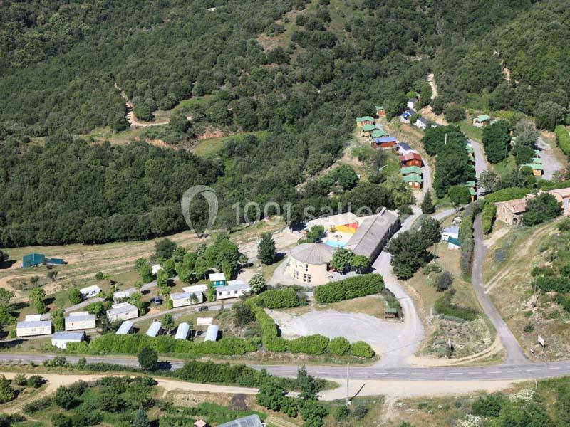 Vue aérienne d'un village entouré de collines boisées, avec des bâtiments, des routes et des espaces verts.