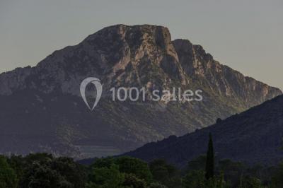 Montagne rocheuse éclairée par une lumière douce, entourée de collines boisées et d'arbres au premier plan.